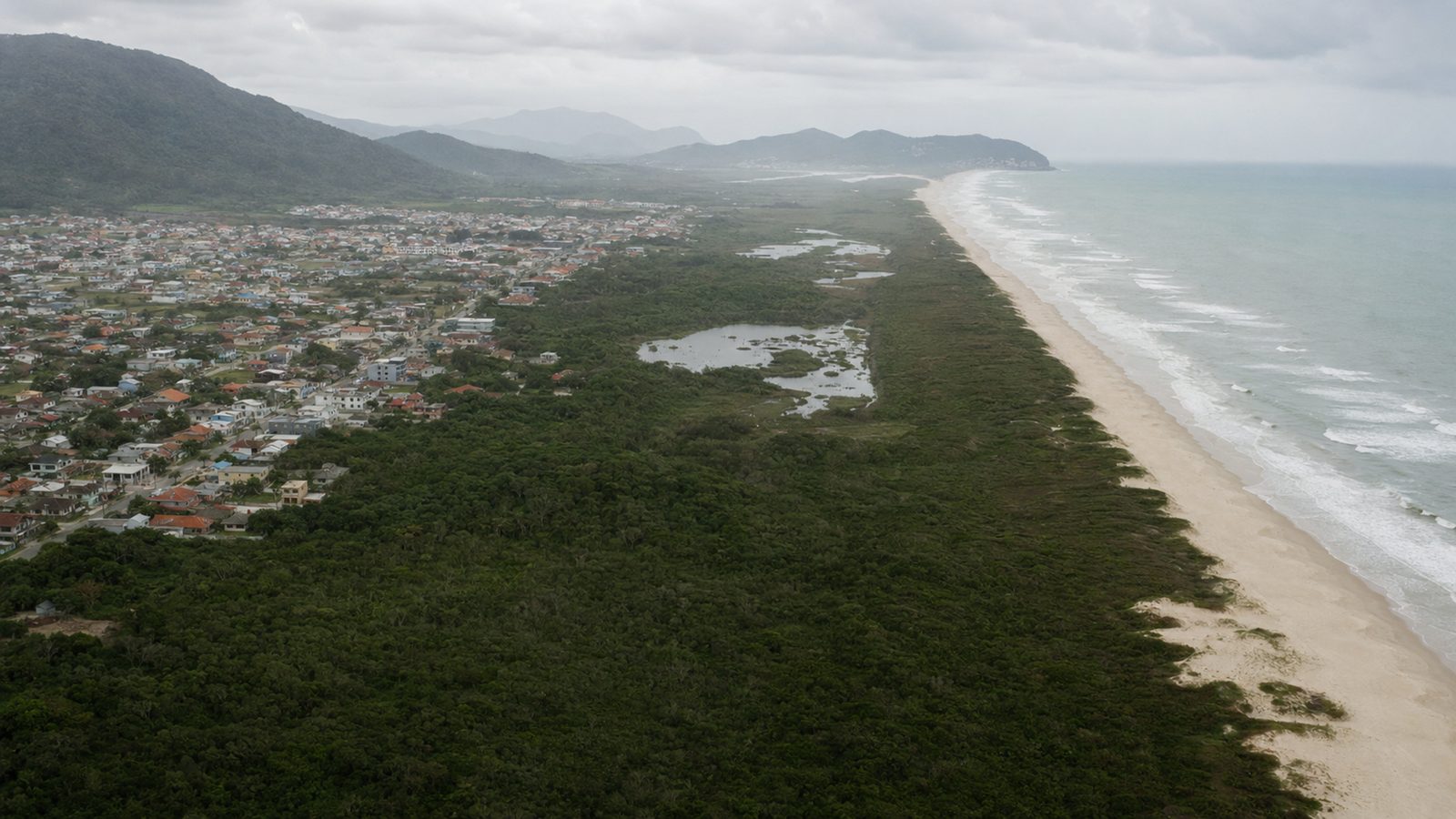 Vista aérea da costa de Santa Catarina: a mancha urbana de um lado, a faixa de restinga e a lagoa preservadas no centro, a praia e o mar do outro — a fronteira que o licenciamento ambiental administra.
