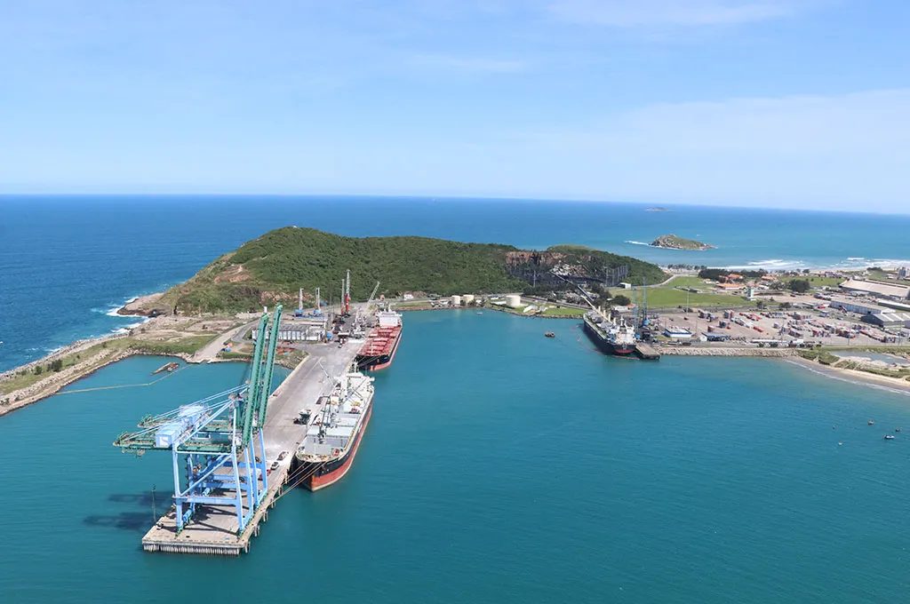 Vista aérea do Porto de Imbituba: guindastes de cais, navios atracados, o molhe e o costão verde ao fundo.